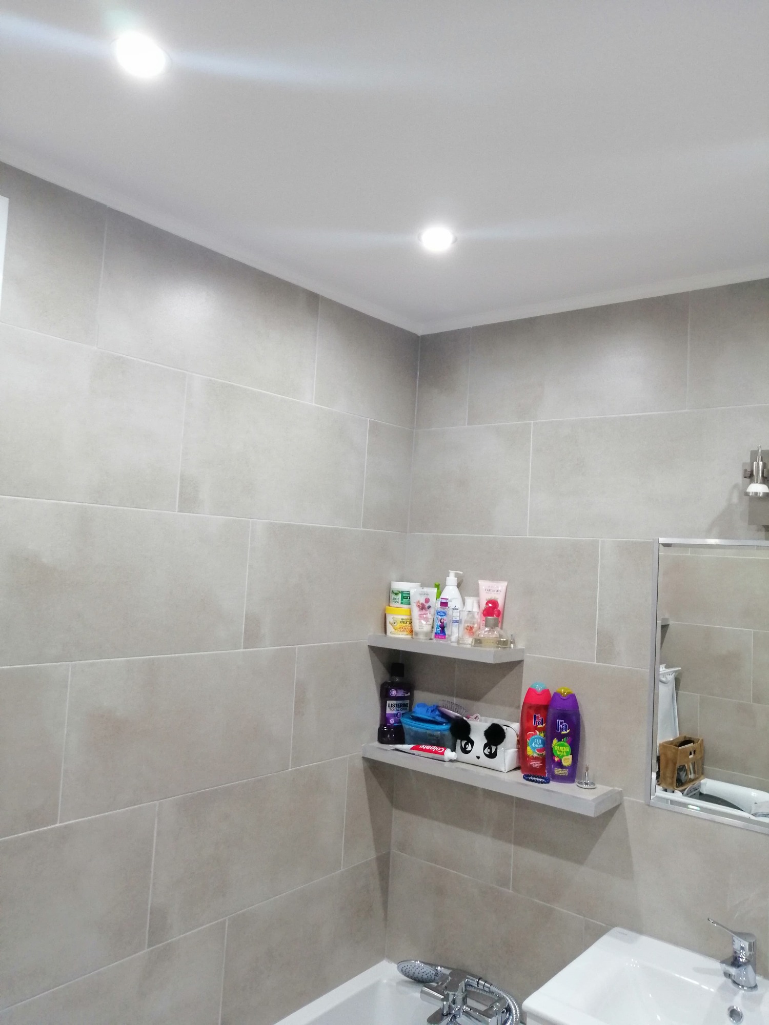 Wykończona łazienka z szarymi, prostokątnymi płytkami, dwiema półkami z kosmetykami i akcesoriami oraz umywalką z baterią.