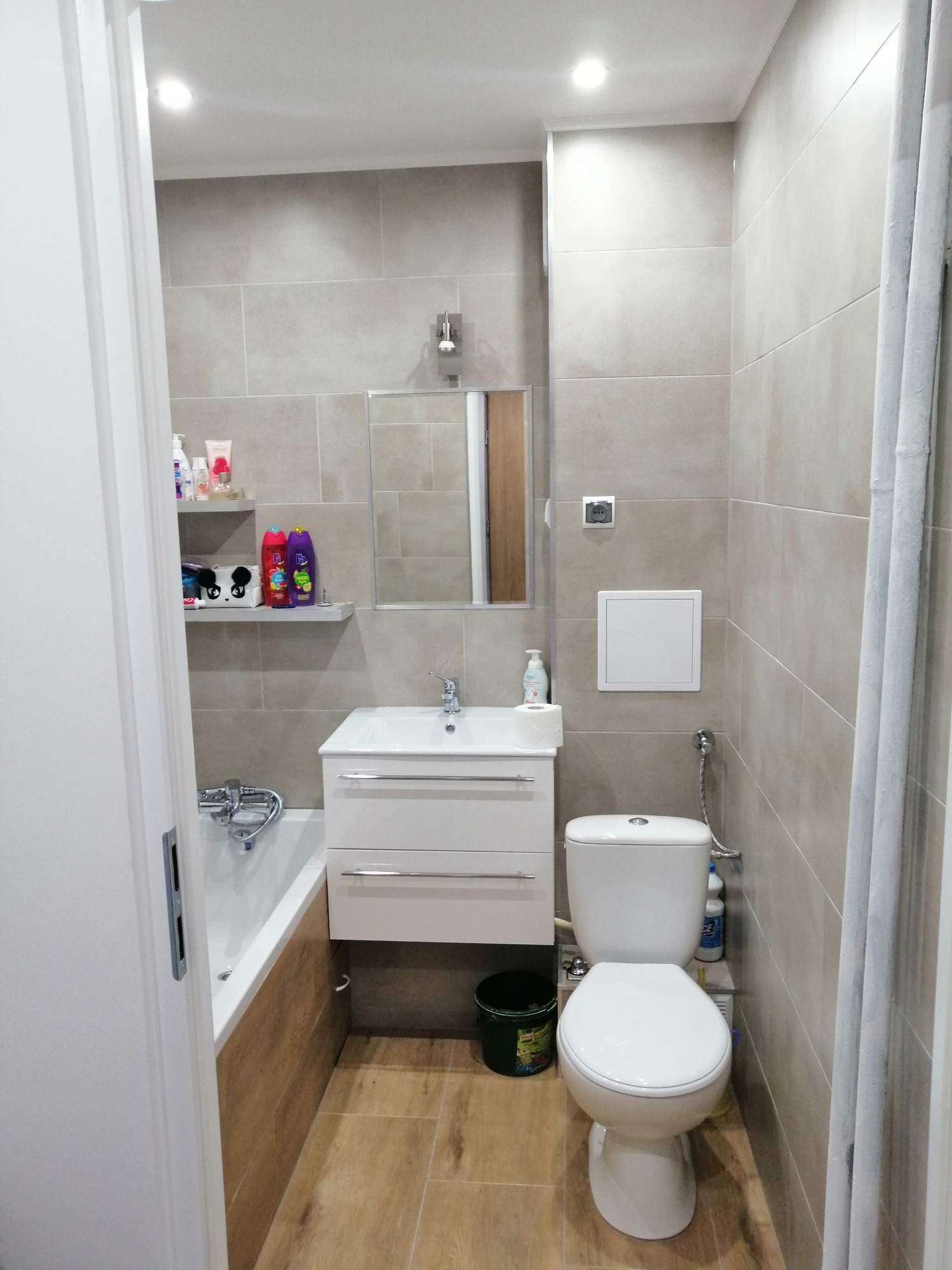 Wykończona łazienka z beżowymi płytkami, białą armaturą, wanną z obudową imitującą drewno i półkami z kosmetykami.