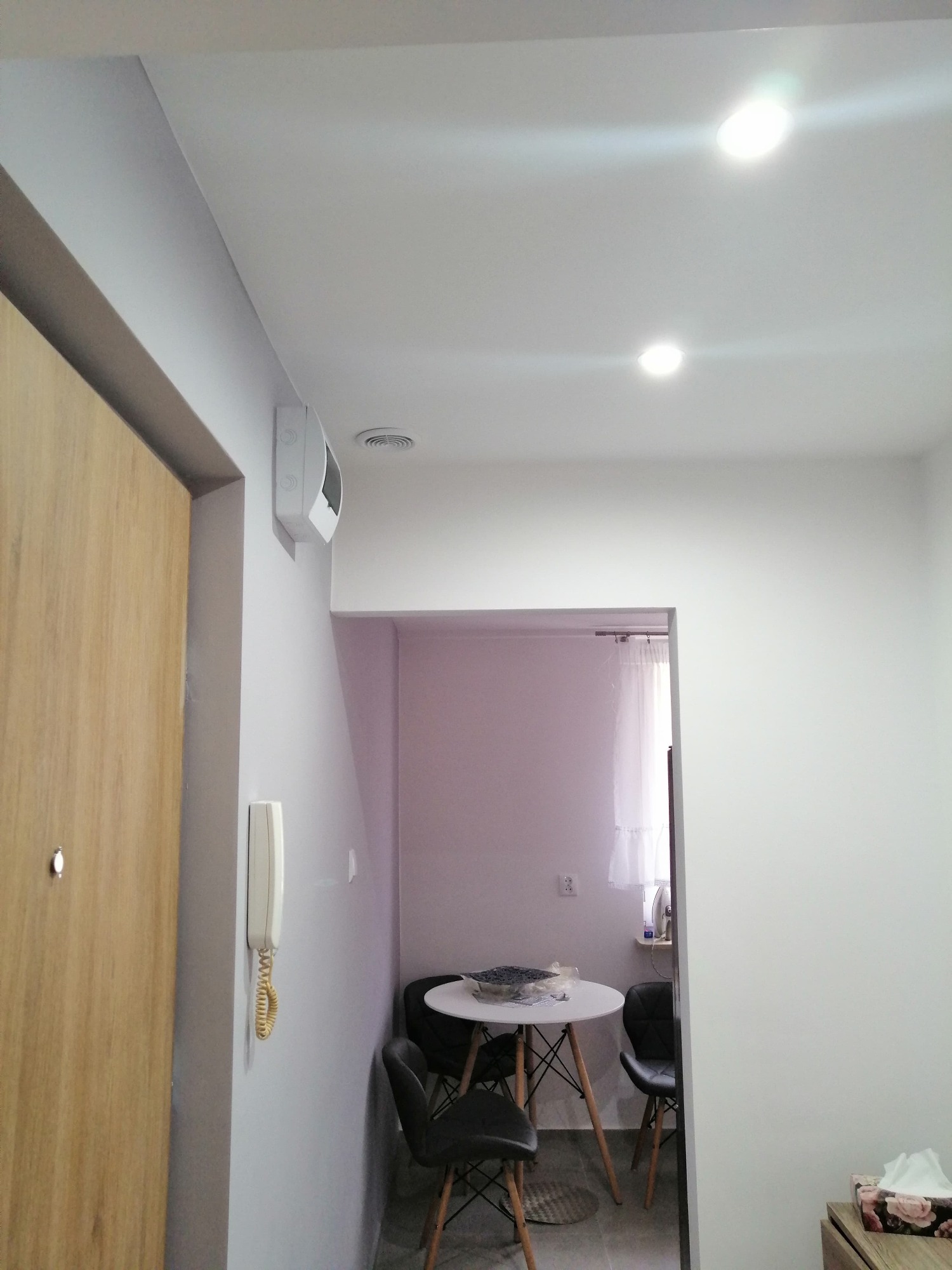 Jasne wnętrze mieszkania po odświeżeniu: pomalowane ściany na szaro, drewniane drzwi wejściowe, biały domofon na ścianie, widoczny fragment jadalni z okrągłym stołem i krzesłami w tle.