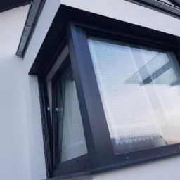 Kątowe okno z ciemną ramą, częściowo otwarte, widoczne z zewnątrz na tle białej ściany budynku z rynną.