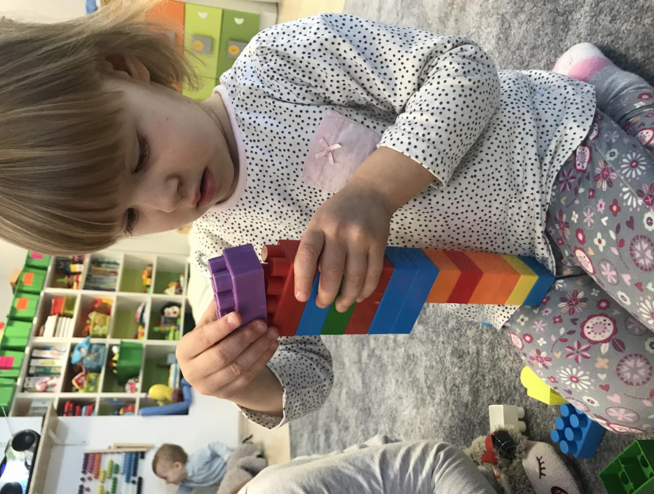 Dziewczynka w bluzce w kropki układa kolorowe klocki na szarym dywanie w pokoju zabaw z regałami pełnymi zabawek w tle.