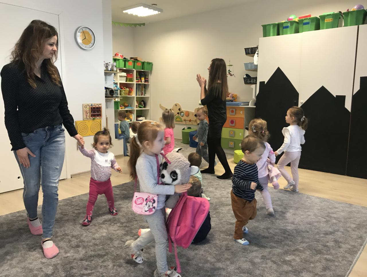 Grupa dzieci w sali zabaw z dwiema opiekunkami, jedna trzyma za rękę małą dziewczynkę, inne dzieci trzymają zabawki, w tle regał z zabawkami i szafa z dekoracją przypominającą sylwetkę miasta.