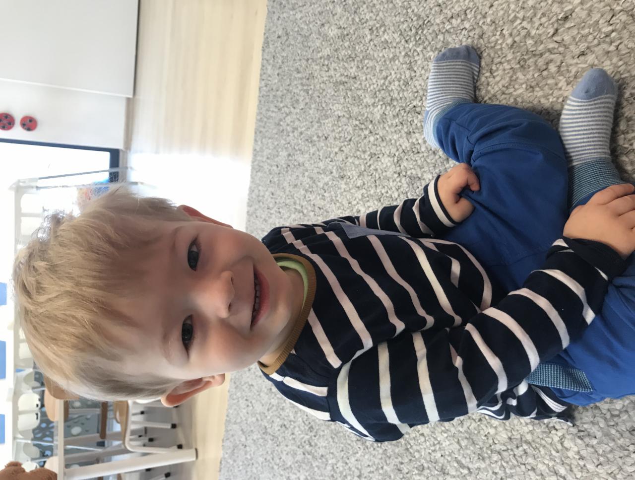 Uśmiechnięty blond chłopiec w paski siedzi na szarym dywanie, w tle widoczne krzesełka dziecięce i okno.