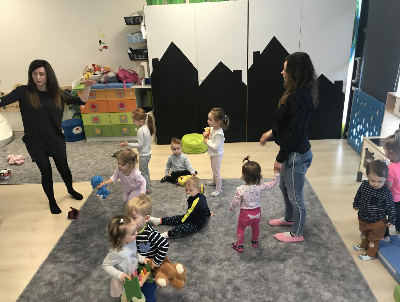 Grupa dzieci w żłobku podczas zajęć ruchowych prowadzonych przez dwie opiekunki, na szarym dywanie, z szafkami w tle.