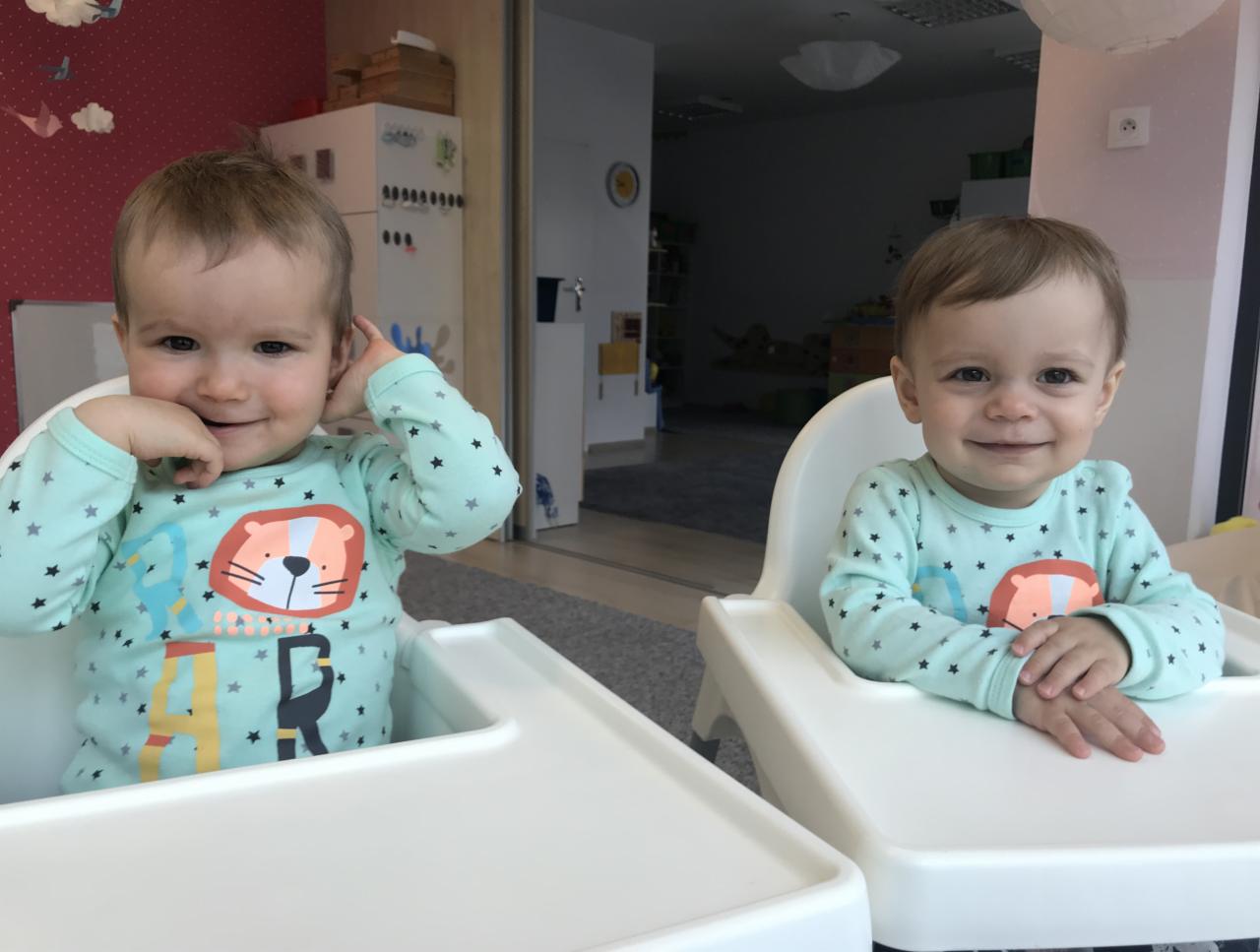 Dwoje uśmiechniętych dzieci w identycznych bluzkach z motywem lwa siedzi w białych krzesełkach do karmienia w żłobku w Lublinie.