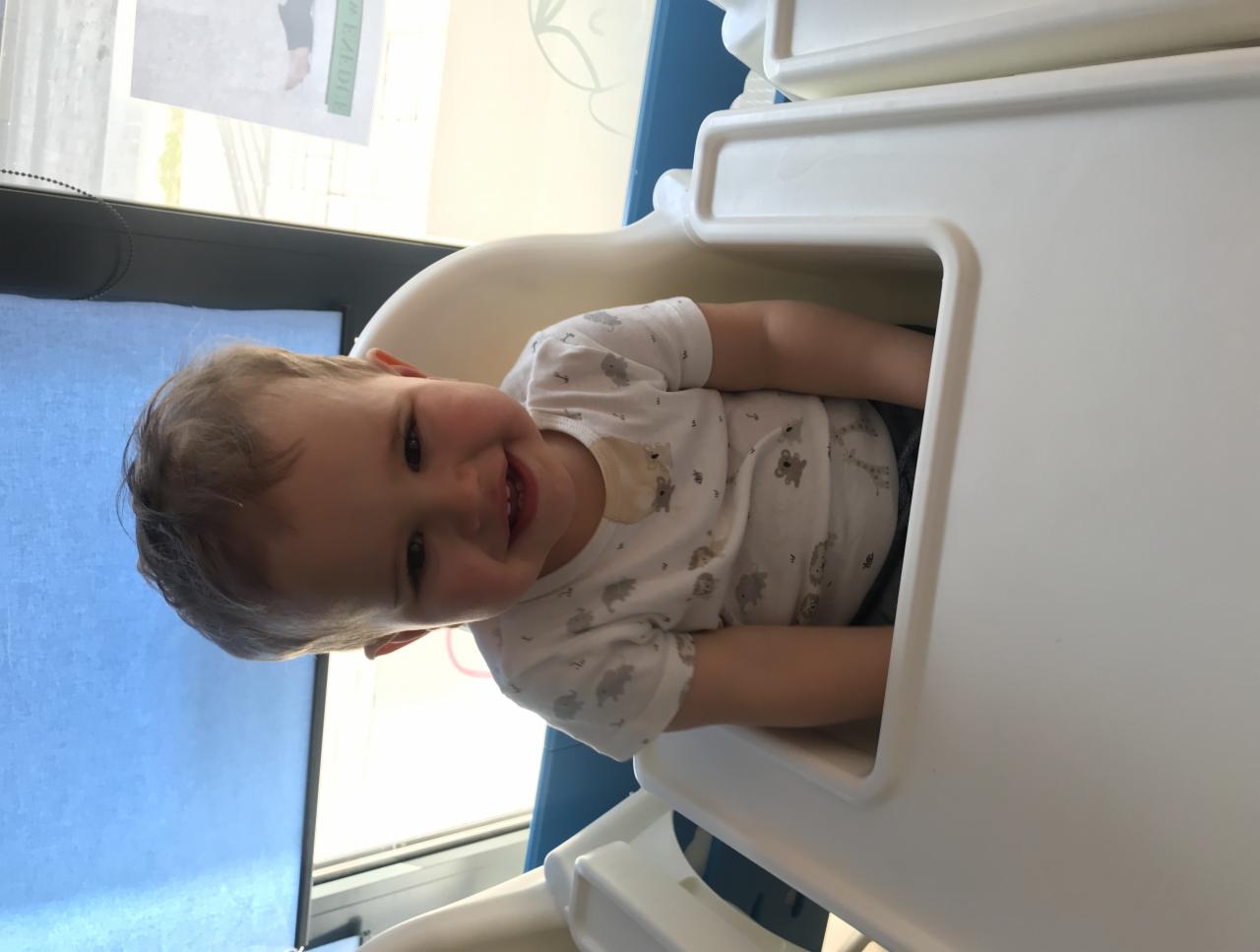 Uśmiechnięty malec w białym body z nadrukiem zwierząt siedzi w białym krzesełku do karmienia w jasnym otoczeniu.