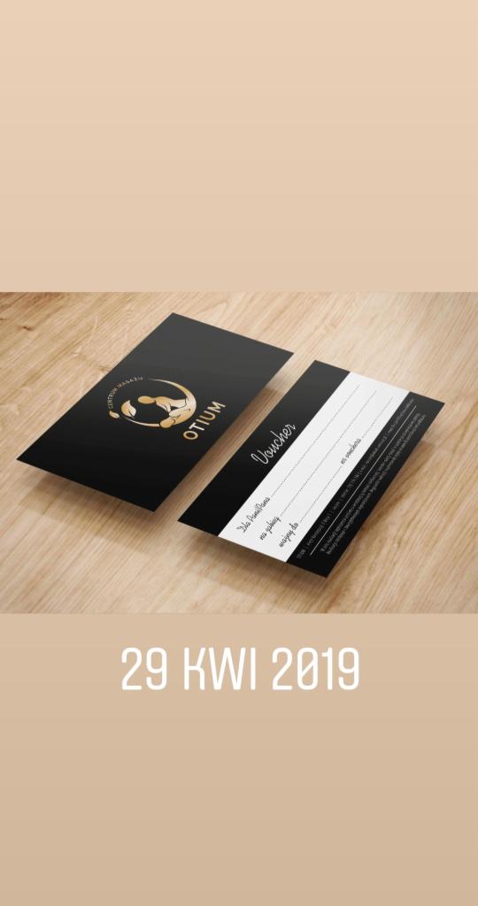 Czarno-złoty voucher na usługi Otium leżący na drewnianym stole. Na voucherze widnieje logo firmy.