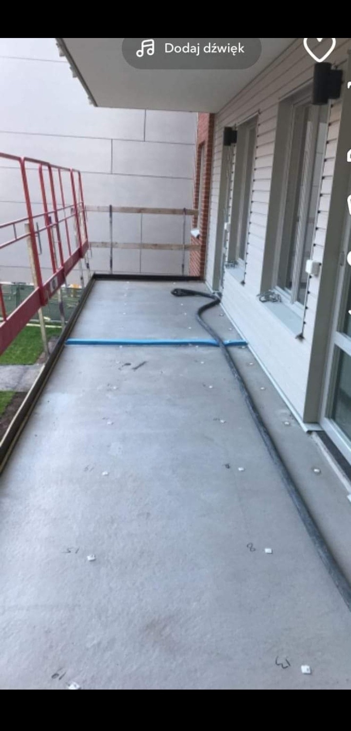 Betonowa posadzka balkonu w trakcie wylewania, widoczna szara wylewka, niebieski wąż do rozprowadzania betonu i rusztowanie ochronne w tle.