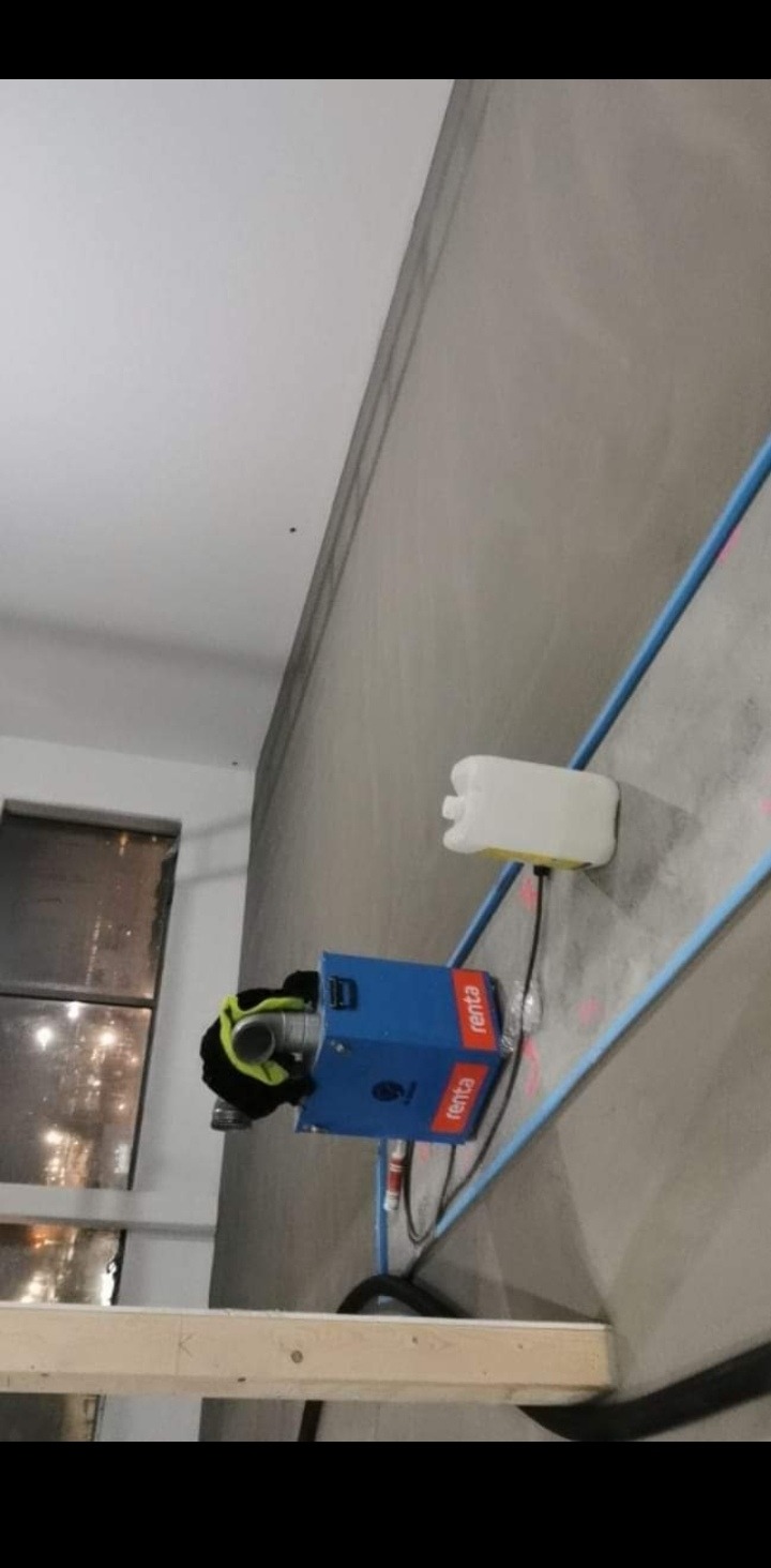 Praca przygotowawcza do remontu biura: Urządzenie do osuszania betonu marki Renta, ustawione w rogu pomieszczenia z surowymi ścianami, widoczne okno i drewniana konstrukcja zabezpieczająca.