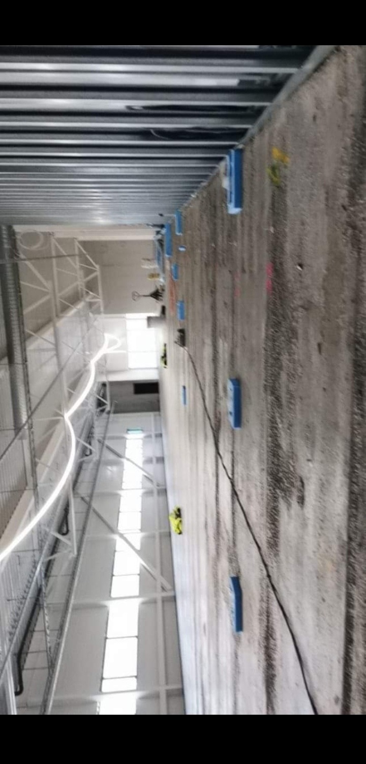 Betonowa ściana w trakcie prac instalacyjnych wewnątrz hali, widoczne niebieskie elementy mocujące i przewody, metalowa konstrukcja sufitu.