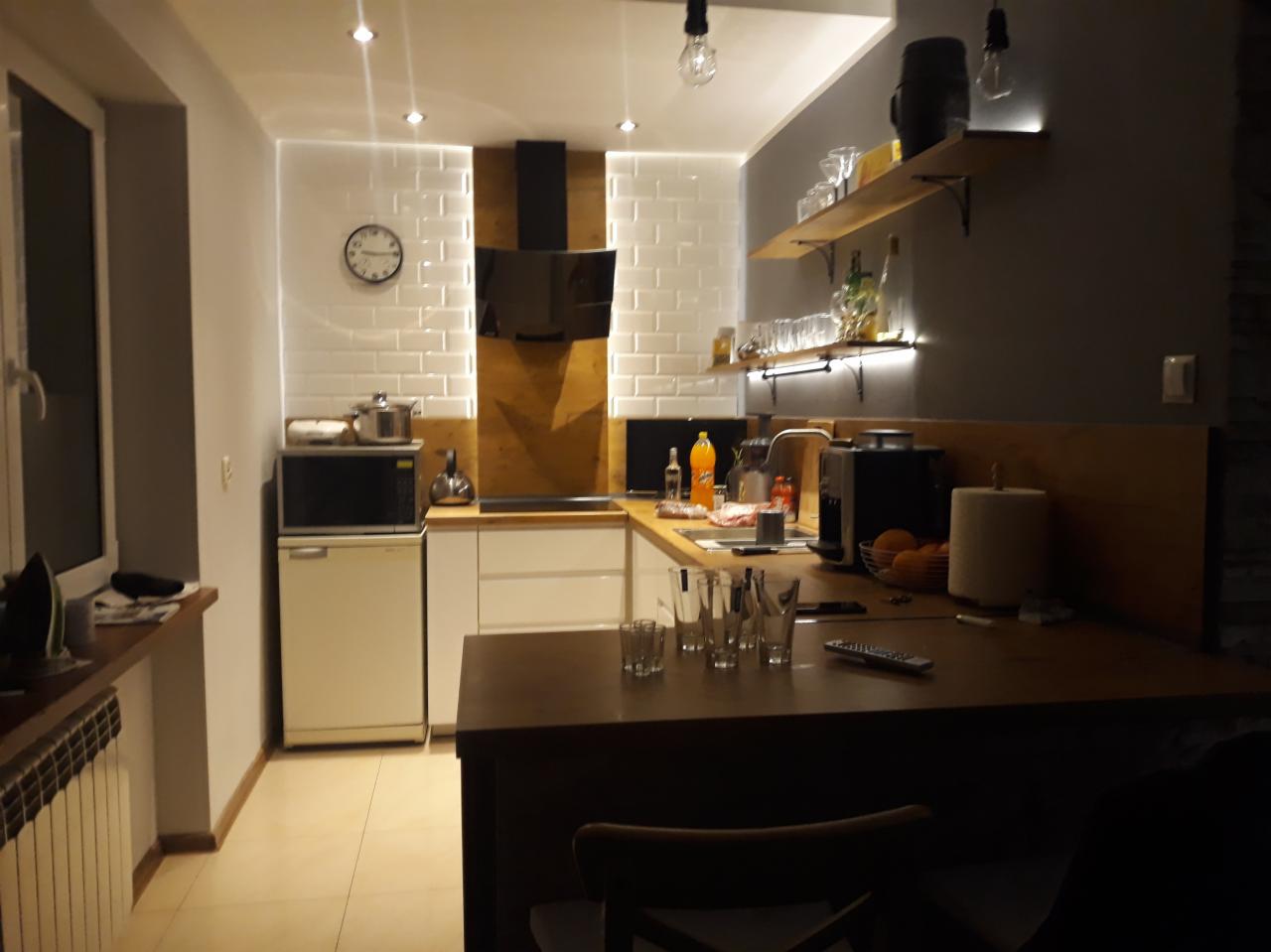 Nowoczesna kuchnia z białymi płytkami imitującymi cegłę, drewnianymi akcentami, podświetlanymi półkami i barkiem z szklankami.