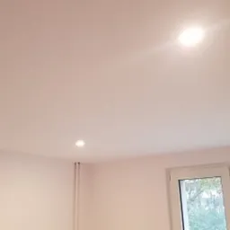 Biały sufit z trzema punktowymi lampami LED, widok na okno z zielenią za szybą.