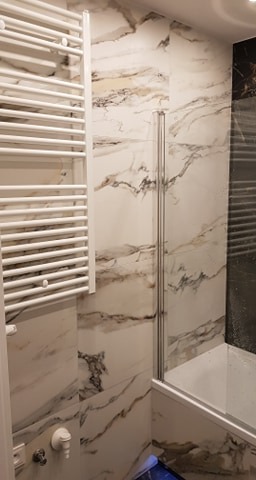 Biała łazienka z marmurowymi płytkami na ścianach, grzejnik drabinkowy i szklany parawan nawannowy.