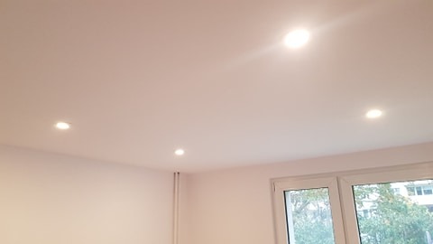 Biały sufit z trzema punktowymi lampami LED, widok na okno z zielenią za szybą.