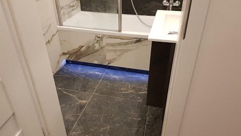 Nowoczesna łazienka z czarną marmurową podłogą i białymi marmurowymi ścianami, oświetlona niebieskim światłem LED przy podłodze, widok zza uchylonych białych drzwi.