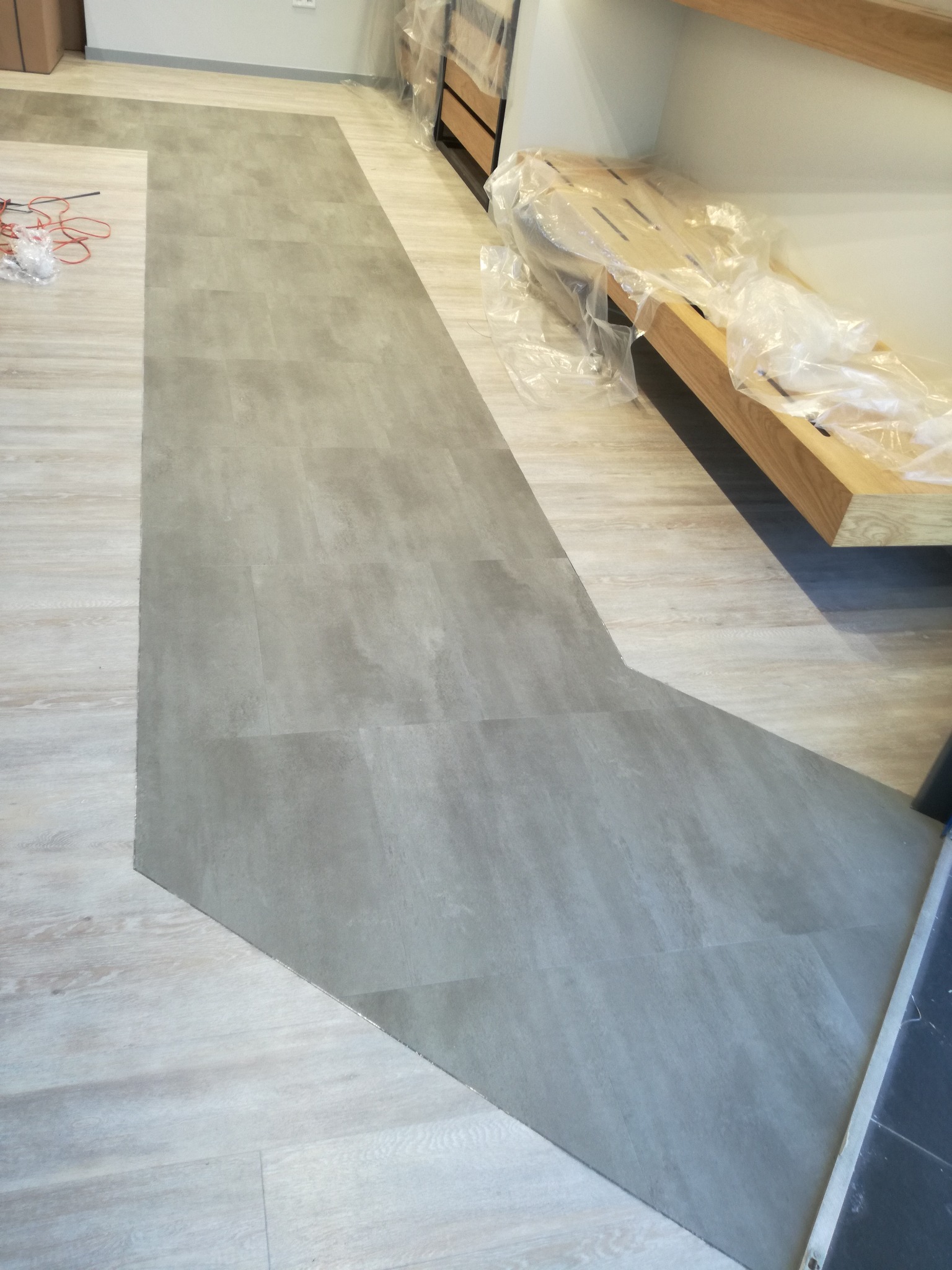 Fragment podłogi z częściowo ułożoną szarą wykładziną imitującą kamień, kontrastującą z jasnymi panelami drewnopodobnymi, widoczne kable i meble zabezpieczone folią ochronną.