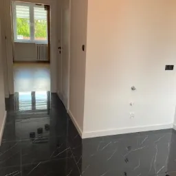 Korytarz z błyszczącą czarną podłogą imitującą marmur, świeżo pomalowane białe ściany, widoczne wyprowadzenia instalacji wodnej i fragment pokoju z oknem.