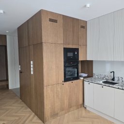 Grupa Investrem - Nowoczesna kuchnia z drewnianą zabudową, zintegrowanym piekarnikiem i mikrofalówką, granitowym blatem, czarnym zlewem i baterią, oraz podłogą w jodełkę.