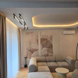 Grupa Investrem - Nowoczesny salon z beżową, modułową sofą, drewnianą podłogą w jodełkę i oświetleniem szynowym. Sufit podwieszany z ciepłą listwą LED, ściana z flizami i grafiką liści.