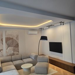 Grupa Investrem - Elegancki salon z beżową sofą modułową, podłogą w jodełkę, podświetlanym sufitem i dekoracyjnymi panelami ściennymi z motywem liści miłorzębu.