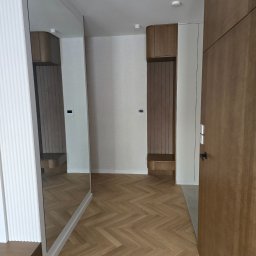 Grupa Investrem - Nowoczesny przedpokój z drewnianą podłogą w jodełkę, dużym lustrem, białymi ryflowanymi panelami i zabudową z drewna. Jasne wnętrze z wbudowaną szafką i siedziskiem.