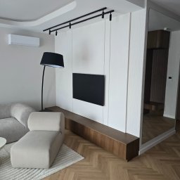 Grupa Investrem - Nowoczesny salon z drewnianą podłogą w jodełkę, białą ścianą TV z ryflowanymi panelami, czarnym oświetleniem szynowym i dużym lustrem. Jasna sofa modułowa, zakrzywiony sufit.