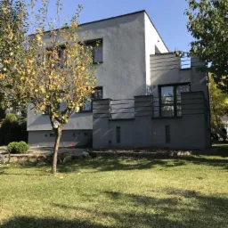 Nowoczesny dom z szarą elewacją, zadbany trawnik i drzewo z żółknącymi liśćmi na pierwszym planie.