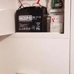 Centrala alarmowa w białej obudowie z akumulatorem MIXPO MI 17-12, czujnikiem dymu z czerwoną diodą i panelem sterowania na ścianie