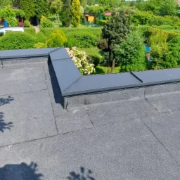 Płaski dach pokryty papą termozgrzewalną z obróbkami blacharskimi w kolorze grafitowym, widok z góry na tle zielonego ogrodu.