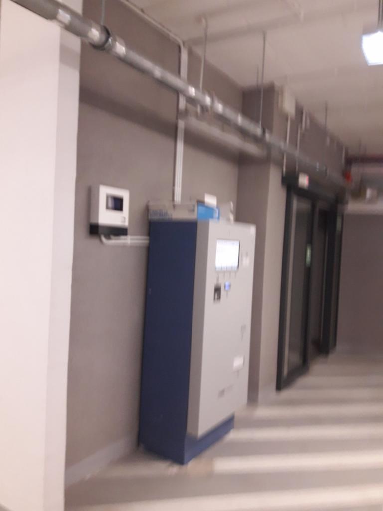 Szara szafa sterownicza z niebieskim dołem i ekranem dotykowym, zamontowana w pomieszczeniu z widocznymi rurami instalacyjnymi pod sufitem i czujnikiem na ścianie.