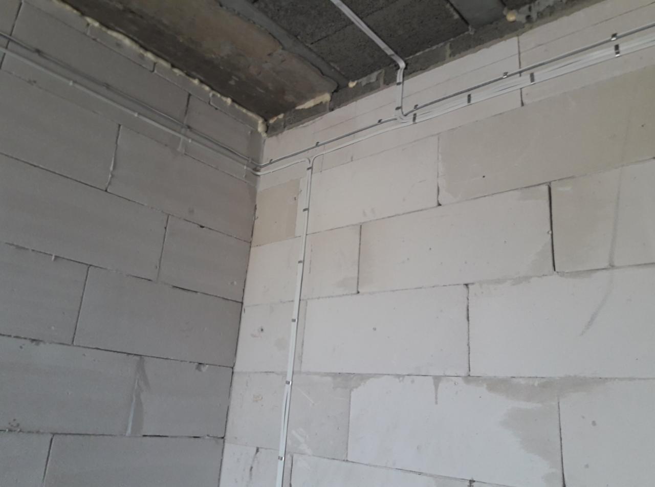 Narożnik surowego wnętrza z widocznymi przewodami elektrycznymi w białych listwach montażowych, poprowadzonymi po ścianach z białych bloczków i szarym, betonowym suficie.