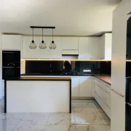 Jasna kuchnia z wyspą, białe fronty szafek, czarny blat i geometryczne lampy wiszące nad wyspą, marmurowe płytki na podłodze.