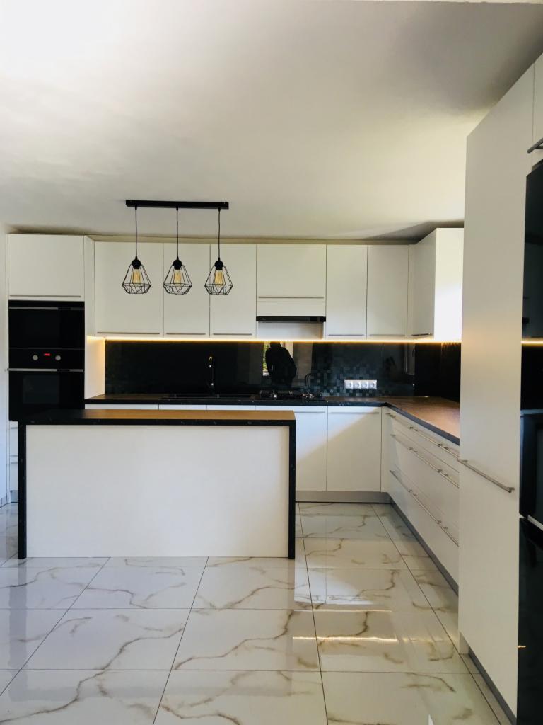 Jasna kuchnia z wyspą, białe fronty szafek, czarny blat i geometryczne lampy wiszące nad wyspą, marmurowe płytki na podłodze.