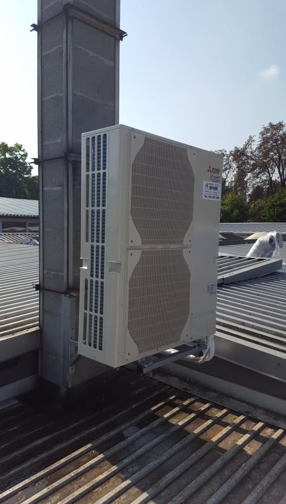 Biała jednostka zewnętrzna klimatyzacji zamontowana na dachu budynku przemysłowego, widoczna instalacja odprowadzająca skropliny.