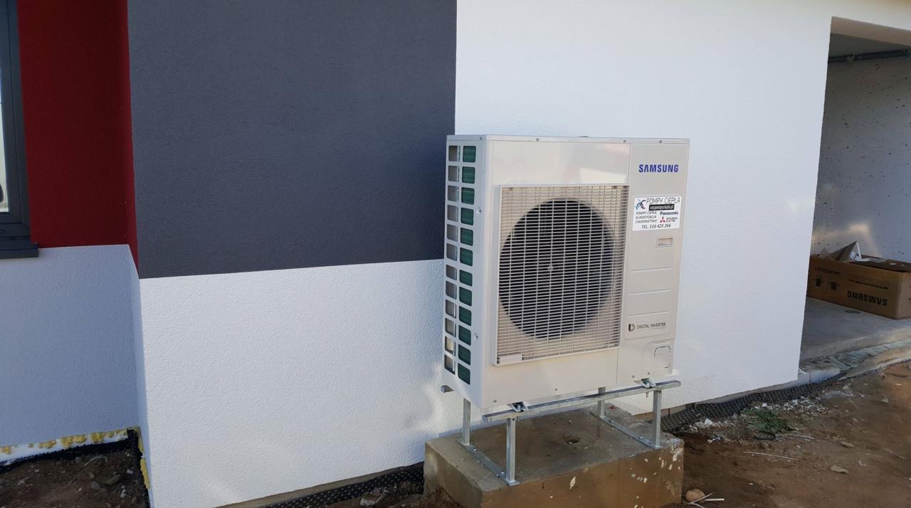 Zewnętrzna jednostka klimatyzacji Samsung Digital Inverter zamontowana na metalowej konstrukcji na betonowym fundamencie przy ścianie budynku z fragmentami elewacji w kolorach czerwonym, szarym...