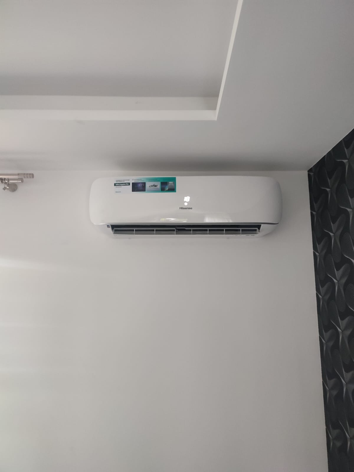 Biały klimatyzator Hisense zamontowany na białej ścianie, widoczny fragment sufitu z wnęką i dekoracyjna tapeta w geometryczny wzór po prawej stronie.