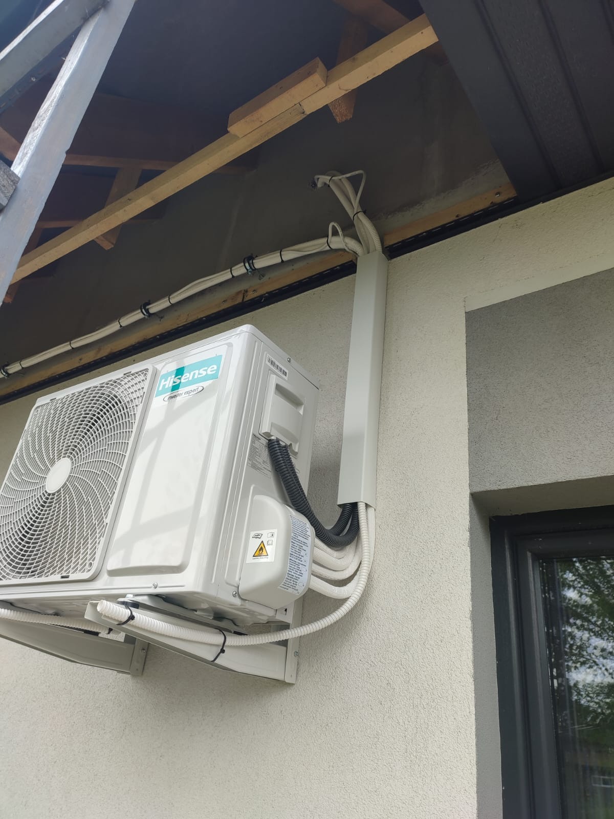 Biała jednostka zewnętrzna klimatyzacji marki Hisense zamontowana na ścianie domu, z widocznymi przewodami w maskownicy i mocowaniami do ściany.