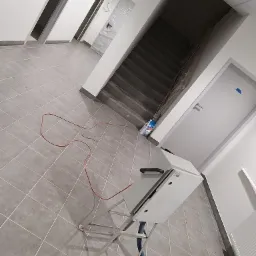 Wnętrze w trakcie prac wykończeniowych: szare płytki podłogowe, białe ściany, schody zabezpieczone folią, oraz maszyna do prac wykończeniowych z niebieskim wężem i czerwonym kablem na podłodze.