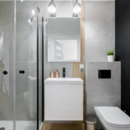 Nowoczesna łazienka z szarą glazurą, kabiną prysznicową z czarnymi detalami, białą szafką z umywalką i lustrem podświetlanym lampami, oraz toaletą z czarnym przyciskiem spłukiwania i czarną ścianą...