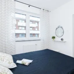 Jasna sypialnia z ciemnoniebieskim łóżkiem, wzorzystymi zasłonami i dekoracyjnym lustrem nad małą półką z zegarem i rośliną.