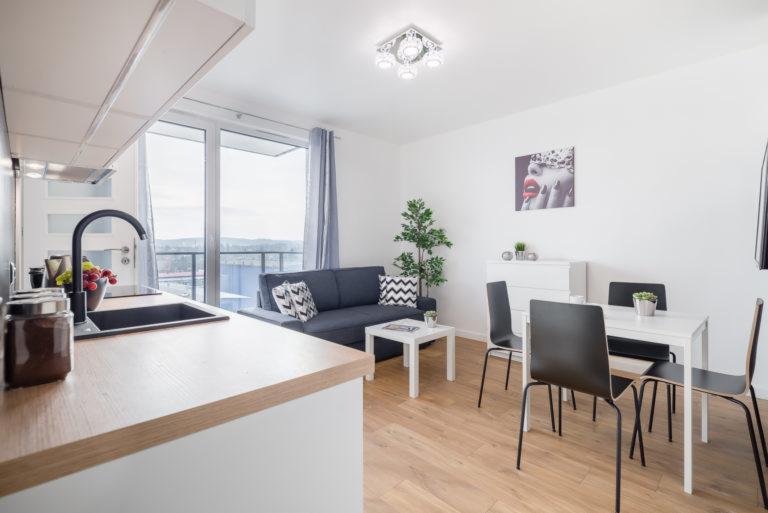 Nowoczesne studio: jasna kuchnia z drewnianym blatem, czarny zlew, widok na salon z szarą sofą i białym stolikiem kawowym, jadalnia z białym stołem i czarnymi krzesłami, parkiet, minimalistyczny...