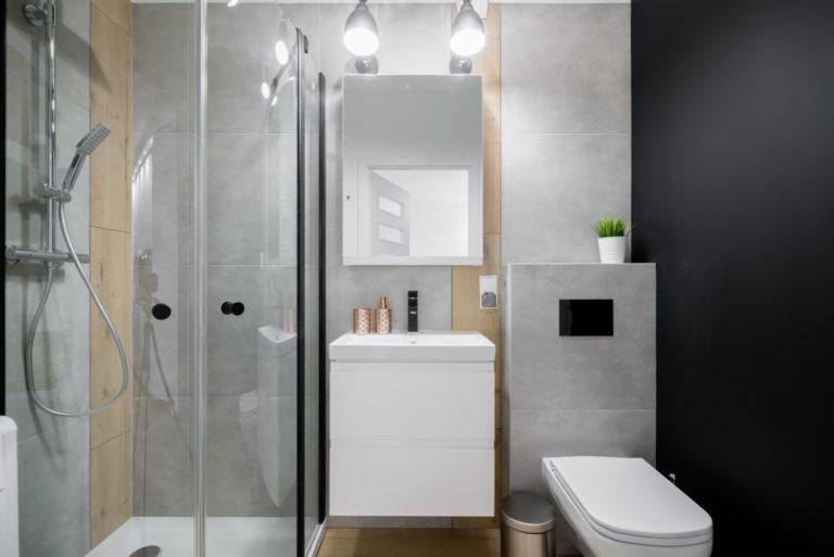 Nowoczesna łazienka z szarą glazurą, kabiną prysznicową z czarnymi detalami, białą szafką z umywalką i lustrem podświetlanym lampami, oraz toaletą z czarnym przyciskiem spłukiwania i czarną ścianą...