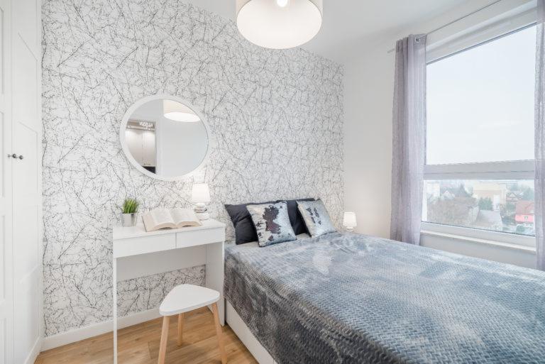 Jasna sypialnia z tapetą w delikatne, abstrakcyjne wzory, białe meble: toaletka z okrągłym lustrem, mała lampka nocna, łóżko z szarą narzutą i dekoracyjnymi poduszkami, drewniana podłoga, duże okno...