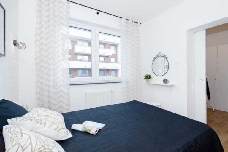 Jasna sypialnia z ciemnoniebieskim łóżkiem, wzorzystymi zasłonami i dekoracyjnym lustrem nad małą półką z zegarem i rośliną.