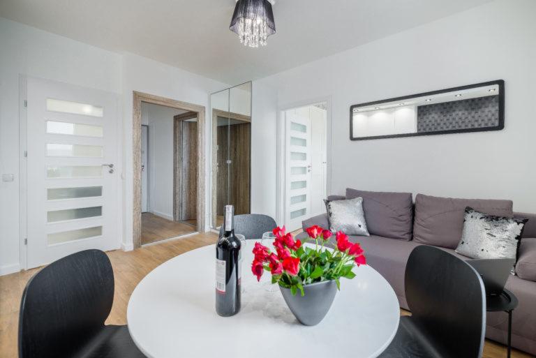 Jasne wnętrze mieszkania z białymi ścianami, okrągłym stołem z czerwonymi różami i butelką wina, szarą sofą z ozdobnymi poduszkami oraz lustrami na ścianie.