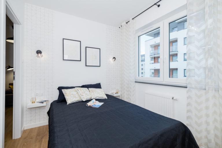 Sypialnia z czarną pikowaną narzutą, wzorzystymi poduszkami i gazetą na łóżku, białe ściany z geometrycznym wzorem, designerskie lampki nocne i minimalistyczne półki, okno z widokiem na nowoczesny...