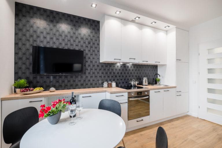 Nowoczesna kuchnia z białymi szafkami, drewnianym blatem, czarną tapetą w romby, telewizorem, okrągłym stołem i czerwonymi kwiatami w wazonie.