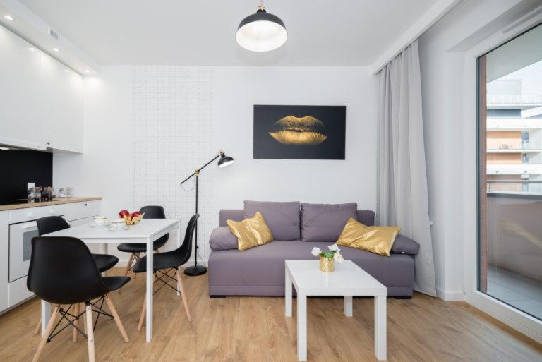Jasne wnętrze mieszkania z otwartą kuchnią, szarą sofą z złotymi poduszkami, białym stołem jadalnym i czarnymi krzesłami, parkiet w ciepłym odcieniu drewna, obraz ze złotymi ustami na ścianie.