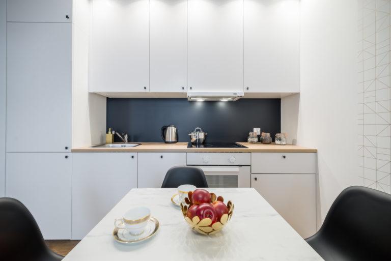 Nowoczesna, minimalistyczna kuchnia z białymi szafkami, czarnym panelem nad blatem, marmurowym stołem i złotymi akcentami w postaci misy z jabłkami i filiżanki.