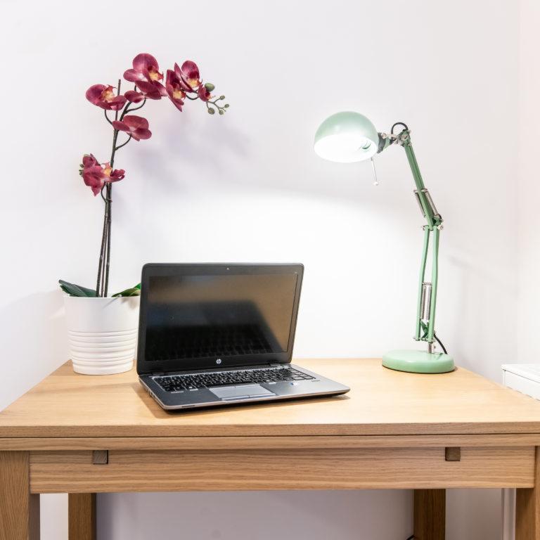 Drewniane biurko z laptopem, zieloną lampką biurkową i storczykiem w białej doniczce na tle białej ściany.